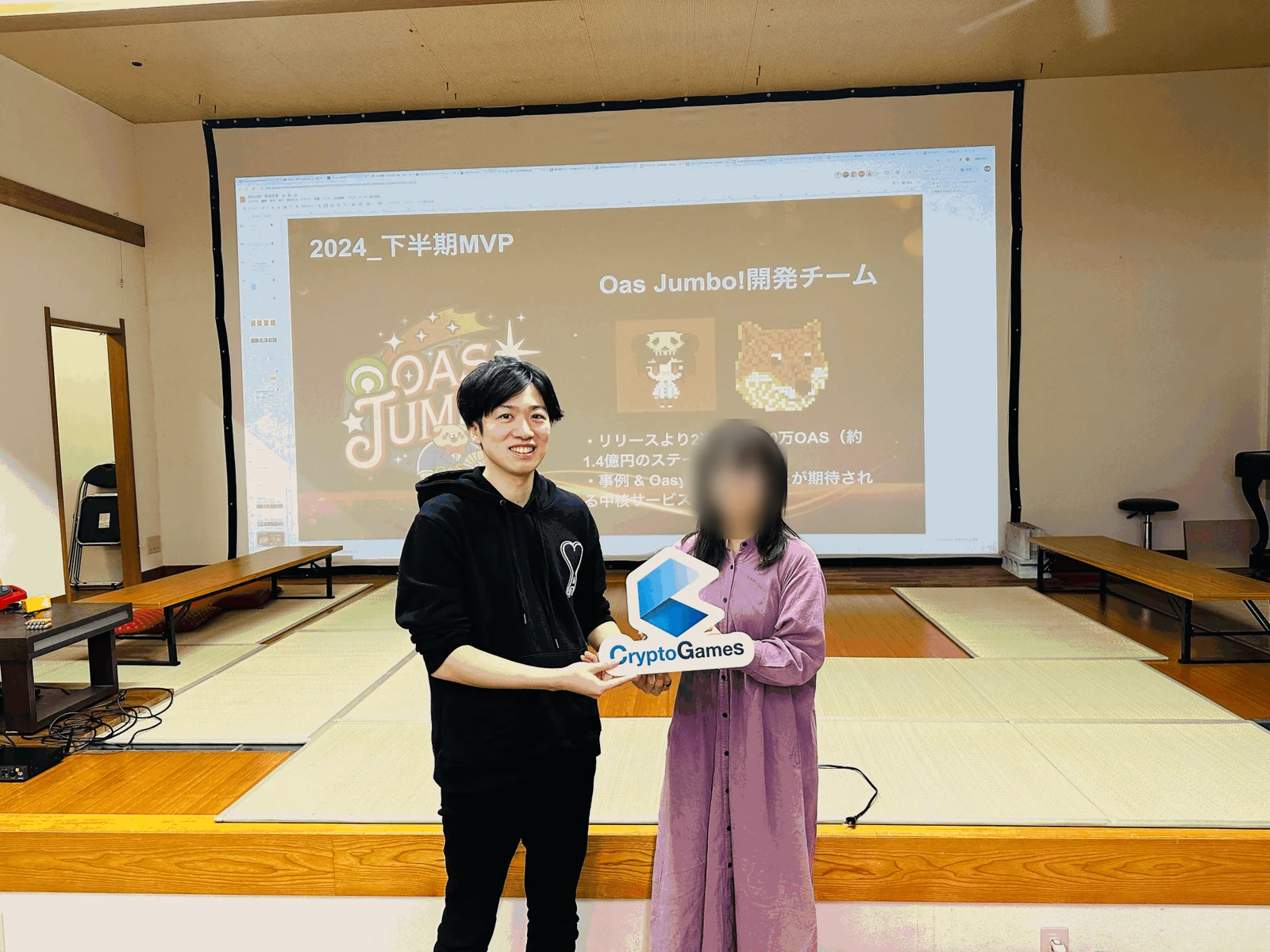 CryptoGamesの社内表彰で、会社のロゴを持つ小澤さん(左)